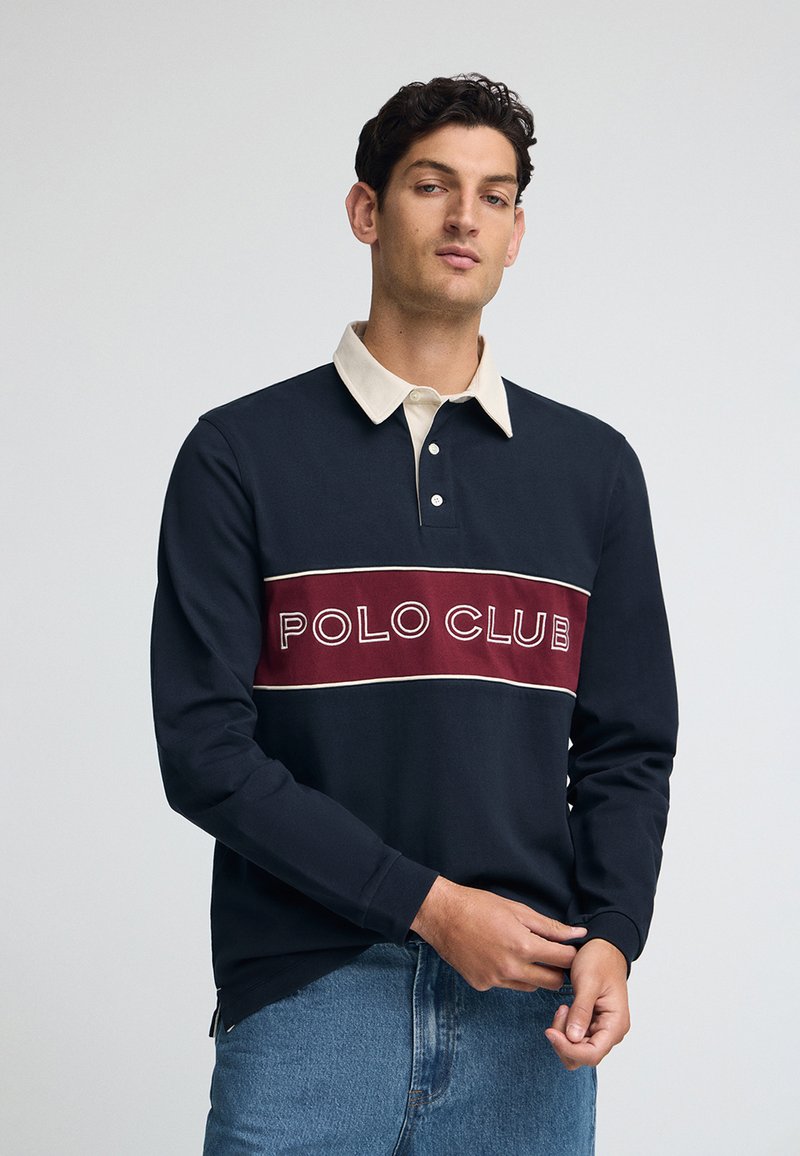 Granatowa koszulka polo z długim rękawem z kremowym kołnierzykiem, z wyrazistym bordowym logiem "POLO CLUB" na piersi, wykonana z bawełnianej tkaniny.
