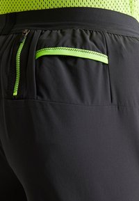 Närbild av svarta träningsbyxor med neongrön dragkedja och trimdetaljer, bärs tillsammans med en neongrön meshtopp.