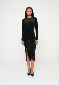 Rochie lungă din catifea neagră cu un design ajustat, având o fante laterală, textură pliată și o finisare netedă. Purtați cu tocuri negre.