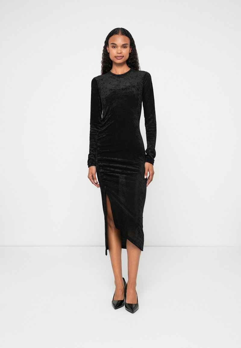 Rochie lungă din catifea neagră cu un design ajustat, având o fante laterală, textură pliată și o finisare netedă. Purtați cu tocuri negre.