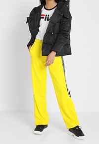 Chaqueta acolchada negra con capucha, camiseta blanca con logotipo rojo y negro, pantalones de chándal amarillos con franjas laterales y zapatillas negras.