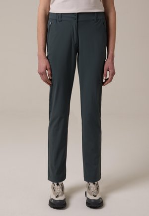 Person steht und trägt dunkelgraue gerade geschnittene Hosen mit Reißverschlusstaschen an den Seiten und beige Wanderschuhe.