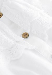 Weiße Baumwollbluse mit aufwendigen Spitzenverzierungen und Holzknöpfen. Texturiert mit sanften, zarten Stickereien.