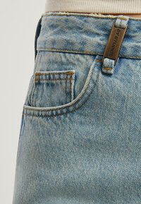 Blugi din denim albastru deschis cu un buzunar frontal mic, cusături contrastante portocalii și un etichetă metalică inscripționată "2Y Studios" pe talie.