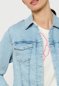 Hele helesinine teksajakk klassikalise kraega, kahe rinnataskuga ja nähtava õmblusega. Allpool valge särk roosa joontega disainiga.