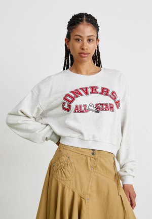 Sudadera corta gris con el texto rojo "CONVERSE ALL STAR" y un gráfico de una zapatilla. Combinada con una falda beige con bolsillos y botones a presión.