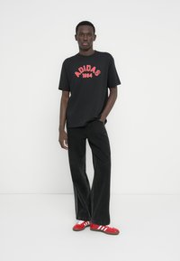 Μαύρο μπλουζάκι με κόκκινο γράφημα "ADIDAS 1994", συνδυασμένο με μαύρο παντελόνι με φαρδιά πόδια και κόκκινα αθλητικά παπούτσια με λευκές ρίγες. Λείο υλικό.