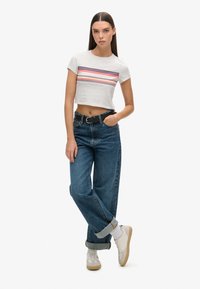 Grijze cropped top met horizontale rode, roze en blauwe strepen, gecombineerd met een high-waisted blauwe jeans en witte sneakers, voorzien van een zwarte riem.