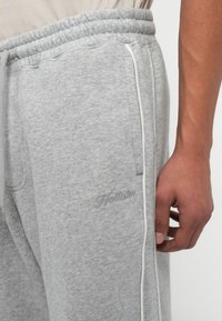 Pelēkie treniņbikses ar elastīgu jostasvietu, baltu auklu, sānu kabatām un izšūtu zīmola logo uz augšstilba. Mīksta tekstūra.