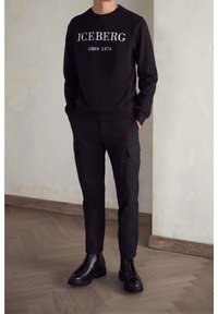 Felpa nera con logo "ICEBERG since 1974", abbinata a pantaloni cargo neri e stivali neri con lacci, in piedi su un pavimento di legno.