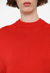 Gros plan du cou et des lèvres portant un pull en maille épaisse rouge vif avec un col côtelé haut.