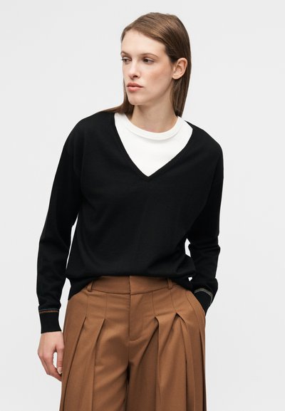 Μαύρο πουλόβερ με V λαιμόκοψη από απαλό ύφασμα, φορεμένο πάνω από λευκή μπλούζα. Συνδυασμένο με ψηλόμεση, καφέ παντελόνια με πιέτες. Διακοσμητικό γαζί στο τελείωμα των μανικιών.