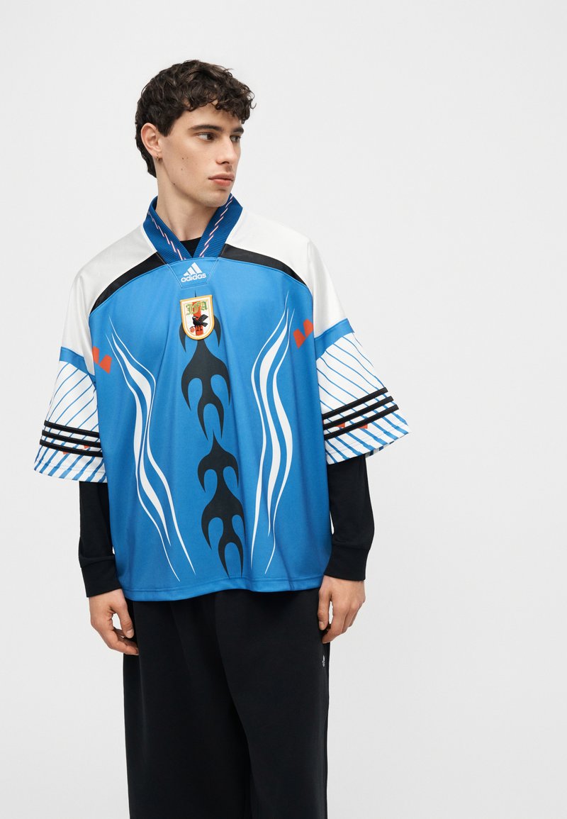 Jeune homme portant un maillot de football bleu, blanc et noir avec des motifs tribaux et un blason, debout devant un fond blanc uni.