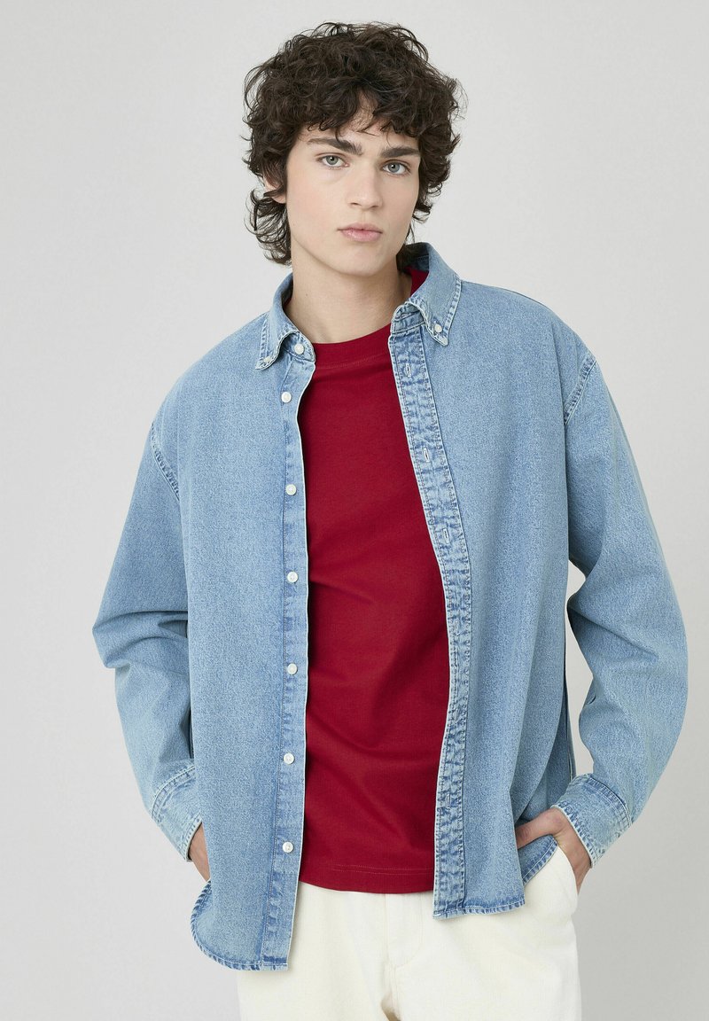 Chemise en denim bleu clair avec fermeture à boutons, col pointu et manches longues portée par-dessus un t-shirt rouge. Texture unie, coupe décontractée.