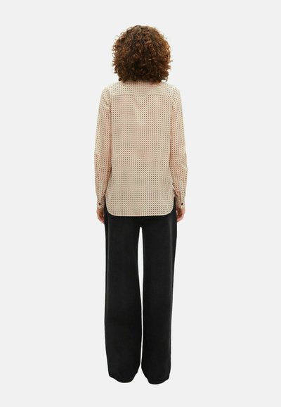 Persona con capelli ricci che indossa una camicia beige a maniche lunghe con pois neri e pantaloni neri larghi, in piedi di spalle su uno sfondo bianco.