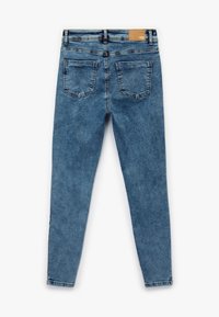 Vaqueros de denim azul de corte ajustado con una textura sutil, cinco bolsillos y un parche de cuero marrón en la cinturilla. Vista trasera mostrada.