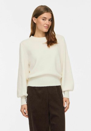Femme portant un pull blanc ample avec de longues manches bouffantes et un pantalon marron foncé taille haute sur fond blanc.