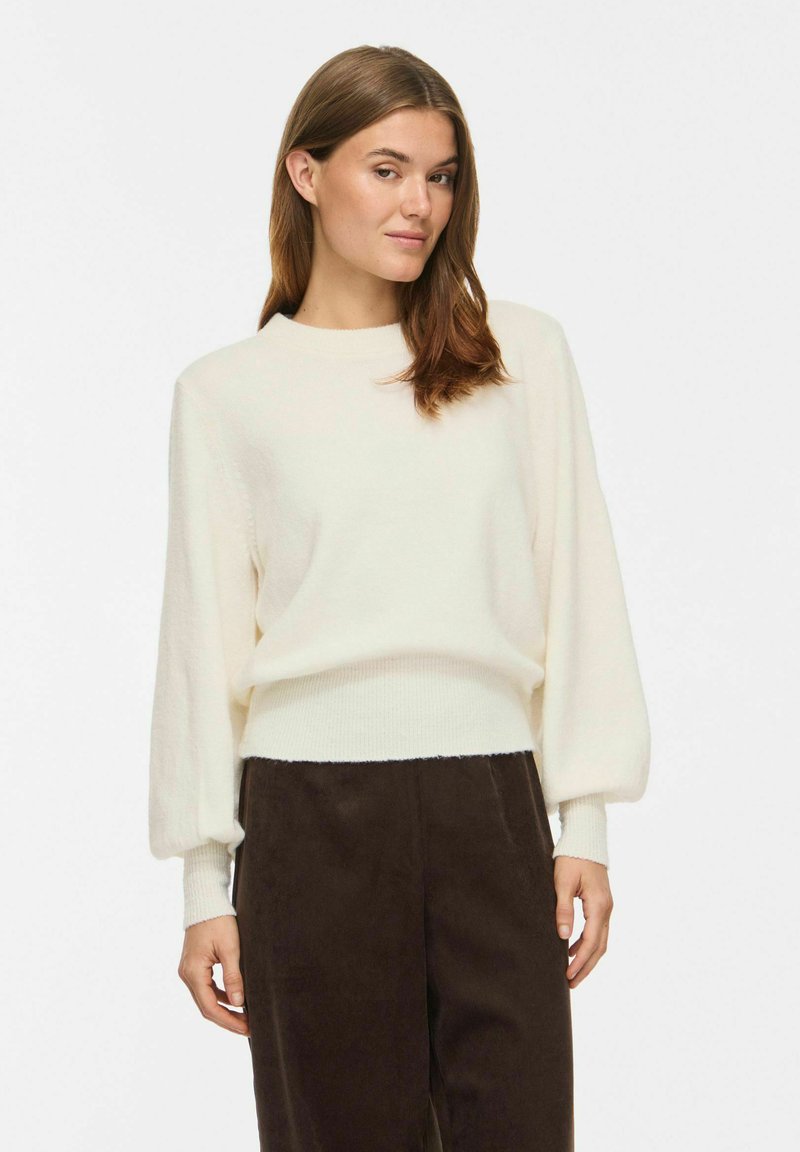 Femme portant un pull blanc ample avec de longues manches bouffantes et un pantalon marron foncé taille haute sur fond blanc.