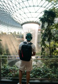 Sac à dos noir avec une tirette rouge, porté par une personne en chemise blanche et en short, surplombant une cascade et une végétation luxuriante sous un dôme de verre.