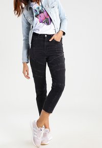 Schwarze Jeanshose mit engem, verkürztem Schnitt und markanten Ziernähten. Kombiniert mit einem farbenfrohen T-Shirt mit Grafik und einer hellen Jeansjacke.