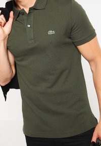 Man som bär en olivgrön Lacoste polo-shirt och håller en svart jacka över axeln, beskuren vid midjan.