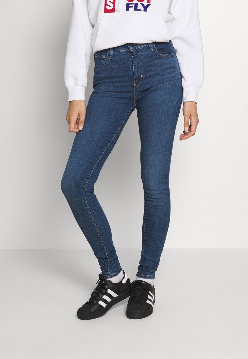 Jeans ajustados de color azul, hechos de denim con un lavado medio. Presentan un bolsillo delantero, trabillas para el cinturón y detalles de costura. Llevados con zapatillas negras.