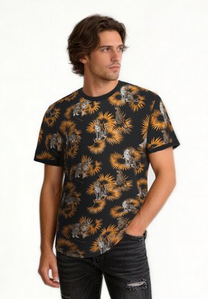 Homme aux cheveux bruns ondulés portant un t-shirt noir avec des motifs orange de palmiers et de léopards, associé à un jean noir déchiré, debout contre un mur blanc.