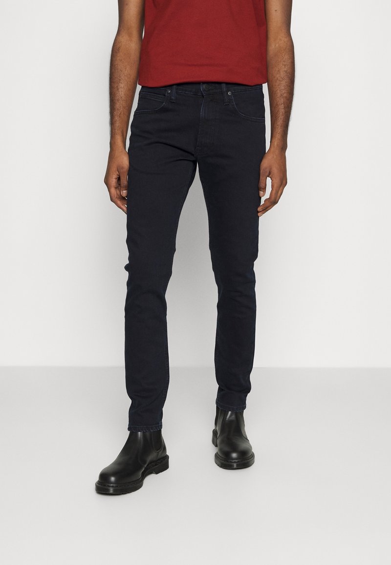 Homme portant un jean foncé ajusté, des bottines en cuir noires et une chemise rouge à manches courtes, debout devant un fond clair uni.