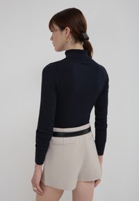 Femme aux cheveux bruns en queue de cheval portant des boucles d'oreilles en or, un pull à col roulé noir, un short beige et une ceinture noire, tournée vers la gauche.