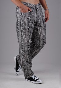 Man wearing black and white patterned pants with hand in pocket and black sneakers with white laces and soles.