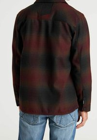 Camicia a quadri in rosso scuro e nero, con una texture morbida, maniche lunghe, polsini con bottoni e dettaglio con giogaia sul retro. Abbinata a jeans blu.