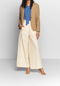 Blazer di lino beige sopra una blusa blu con dettaglio al collo annodato, abbinato a pantaloni a gamba larga crema e tacchi aperti color beige.