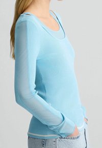 Haut bleu clair à manches longues côtelées avec manches en maille transparente, encolure dégagée et silhouette ajustée, associé à un denim.
