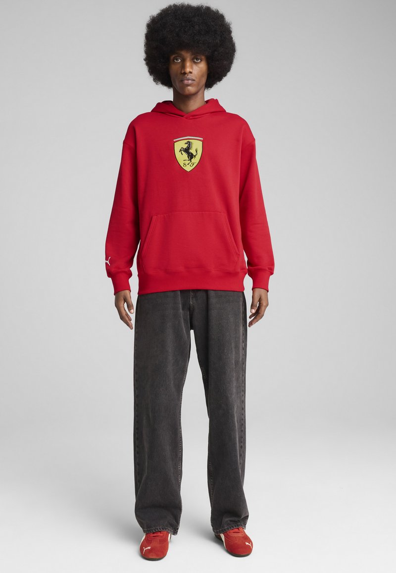 Rød hoodie med et gult Ferrari-logo på brystet, stor forlommer, parret med sorte løse jeans og røde træningssko.