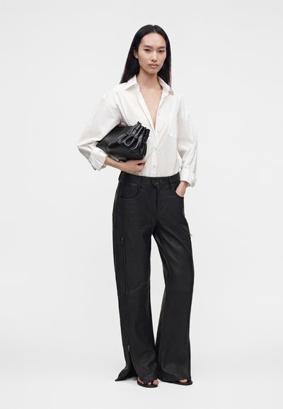 Femme portant une chemise blanche boutonnée et un pantalon en cuir noir à jambes larges, tenant une pochette noire froncée sur fond blanc.