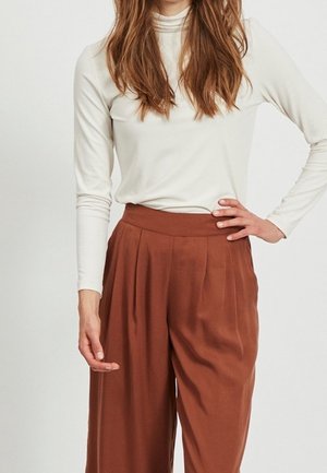 Femme portant un col roulé blanc à manches longues et un pantalon ample taille haute couleur rouille, debout avec une main sur la hanche.