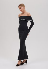 Svart off-shoulder klänning med långa ärmar, utsvängd fåll, vit accentkant och elegant, åtsittande design. Bärs med svarta högklackade skor.