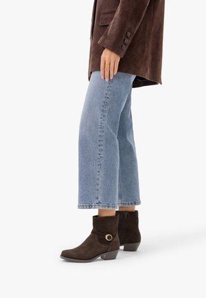 Personne portant un jean court bleu clair, des bottines en daim marron foncé à talons bas avec une boucle et une veste marron foncé.