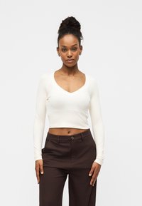 Λευκό crop top με μακριά μανίκια και λαιμόκοψη σε σχήμα V, κατασκευασμένο από απαλό υλικό, συνδυασμένο με καφέ ψηλόμεσα παντελόνια.