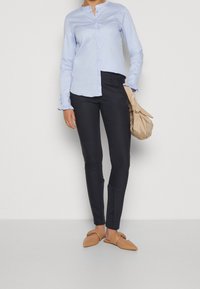 Ljusblå, långärmad skjorta med knappar och volangkantade ärmar, kombinerad med åtsittande mörka byxor och beige platta skor, hållande en beige väska.