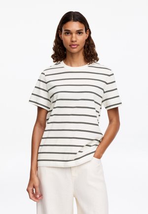 Junge Frau mit lockigem Haar, die ein weißes T-Shirt mit dünnen schwarzen horizontalen Streifen und weiße Hosen trägt und mit einer Hand in der Hosentasche steht.