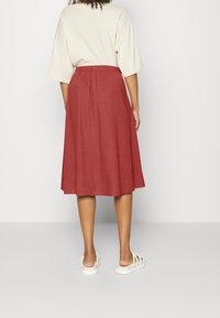 Femme portant une jupe rouge rouille jusqu'au genou, un t-shirt oversize crème et des sandales blanches à glisser, se tenant devant un fond uni clair.