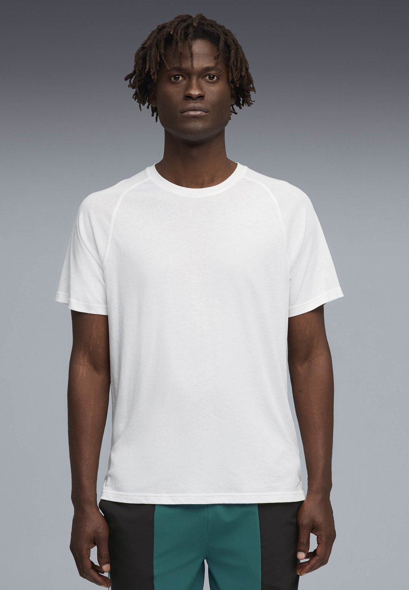 Νεαρός άνδρας με κοντά dreadlocks, ντυμένος με απλό λευκό κοντομάνικο μπλουζάκι και πράσινο-μαύρο παντελόνι, στέκεται μπροστά σε γκρίζο φόντο.