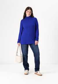 Blå langermet høyhalset topp kombinert med mørke denimjeans. Modellen holder en lys grå håndveske med teksturert stropp og har på seg beige sneakers.