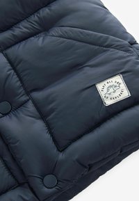 Giacca puffer blu navy con un motivo a rombi, bottoni neri tondi e una toppa di tessuto bianco con un grafico di un animale giocoso.