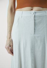 Jupe en coton blanche présentant une coupe droite, des plis sur le devant et une fermeture à boutons. Tissu texturé avec des détails de couture subtils.