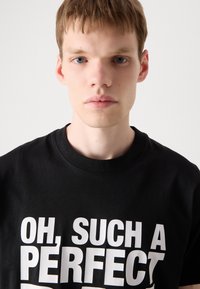Černé bavlněné tričko s výrazným bílým textem, který zní "OH, TAK DOKONALÉ". Klasický design s kulatým výstřihem a krátkými rukávy.