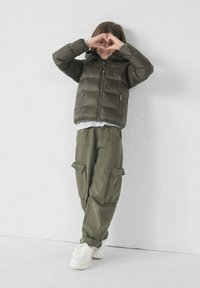 Olijfgroene pufferjack met een capuchon, ritsen en een gestikte afwerking, gecombineerd met olijfkleurige cargobroeken en witte sneakers. Kind vormt een hartvorm.