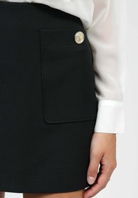 Gros plan sur une jupe noire avec une poche avant et un bouton doré, associée à un poignet de chemise à manches longues blanche et une main visible portant une bague.