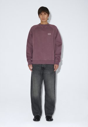 Junge Person steht aufrecht, trägt einen mauvefarbenen Pullover, lockere ausgebleichte schwarze Jeans, schwarze Schuhe und silberne Creolen vor einem einfarbigen Hintergrund.